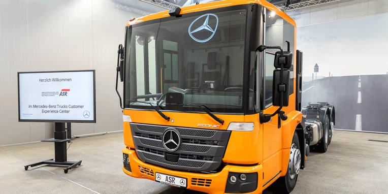 Ein orangefarbener Mercedes-Benz Lkw steht im Mercedes-Benz Trucks Kunden Erlebniszentrum, daneben ein Bildschirm mit Willkommensnachricht und Unternehmenslogo.