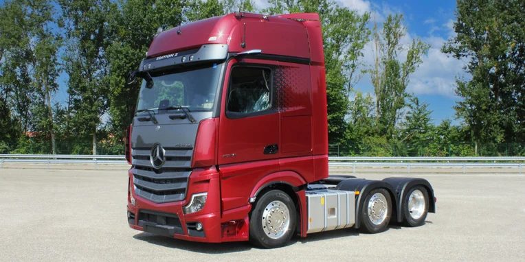 Camion Mercedes-Benz rosso in posizione di profilo, con cabine alta e dettagli moderni, posizionato su una superficie stradale con vegetazione sullo sfondo.