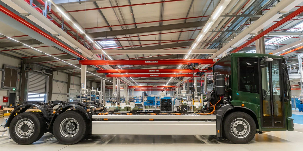 Ein Lkw-Chassis steht in einer modernen Fabrikhalle. Der Lkw ist grün lackiert und hat keine Aufbauten. Im Hintergrund sind Fertigungsmaschinen und -anlagen sichtbar, die für die Montage genutzt werden. Die hohe Decke und das industrielle Ambiente sind erkennbar.