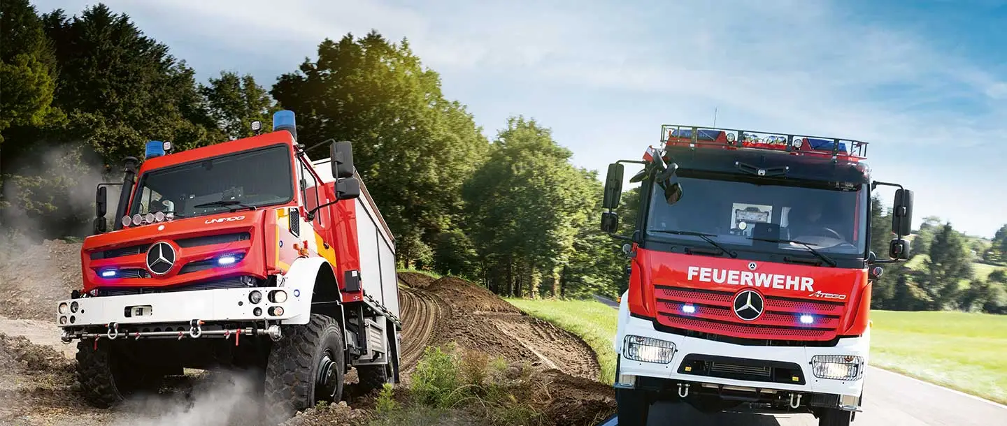 Zwei Feuerwehrfahrzeuge von Mercedes-Benz stehen auf unbefestigtem Gelände und einer Straße. Die Fahrzeuge sind rot lackiert und mit Blaulicht ausgestattet, bereit für Einsätze.