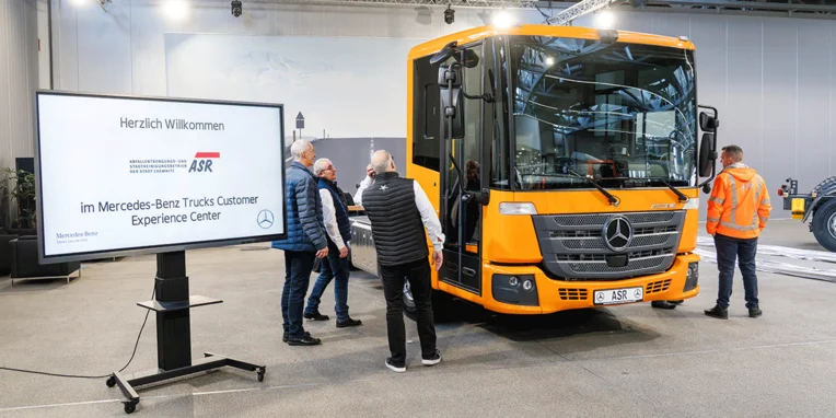 Willkommenden Personen stehen um einen gelben Mercedes-Benz Lkw herum, während eine digitale Anzeigetafel Informationen zum Mercedes-Benz Trucks Customer Experience Center zeigt.
