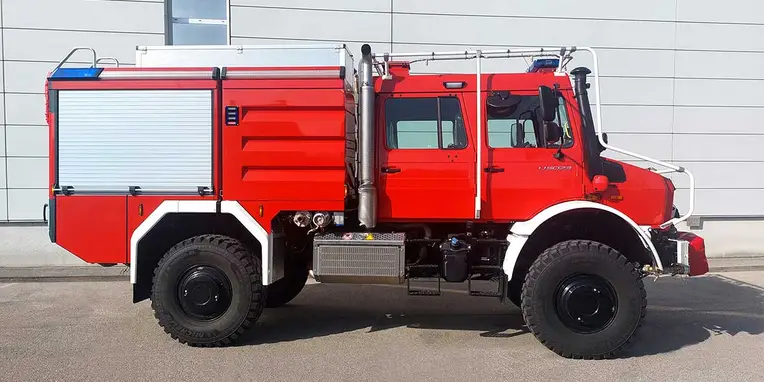 Roter Feuerwehr-Lkw mit großen Reifen, ausgestattet mit einem Wassertank und verschiedenen Ablagen, steht an einer Wand mit grauer Verkleidung.