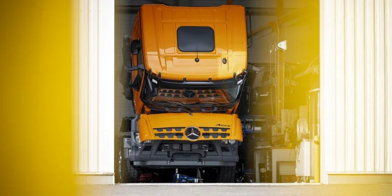 Camion Mercedes-Benz arancione parcheggiato in un garage, con il cofano sollevato. L'immagine mette in evidenza la robustezza e la funzionalità del veicolo commerciale.