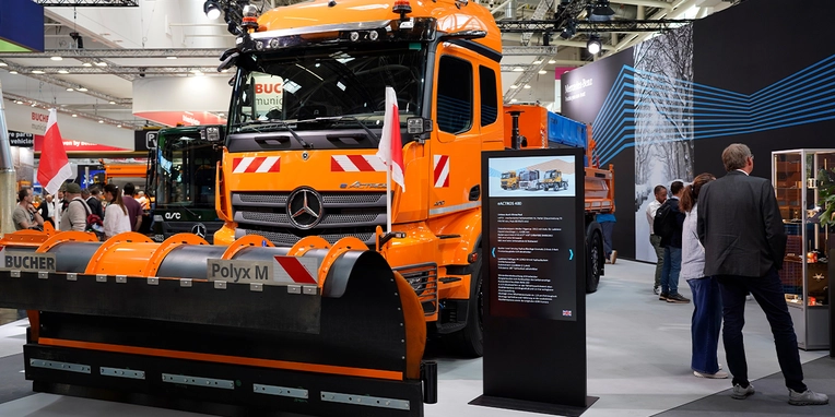 Schnee- und Räumfahrzeug von Mercedes-Benz mit einem großen Schneepflug auf einer Messe, umgeben von Besuchern und Informationsständen zu Dienstleistungen und Produkten.