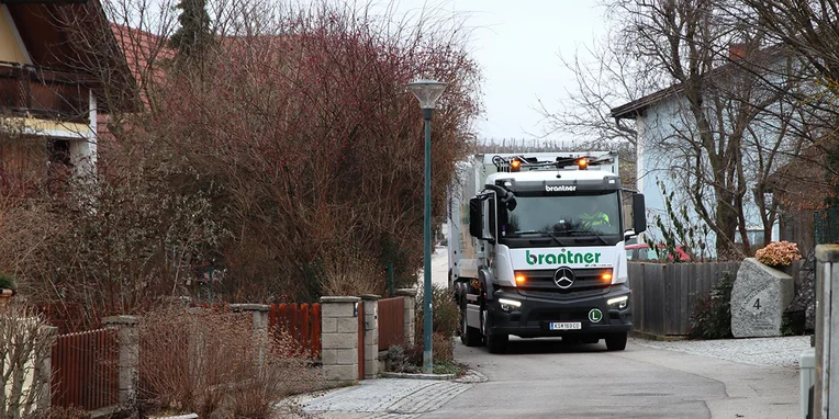Ein Mercedes-Benz Lkw fährt in eine schmale Straße in einem Wohngebiet, während sich Bäume und Häuser in der Umgebung befinden. Der Lkw trägt die Aufschrift "brantner".