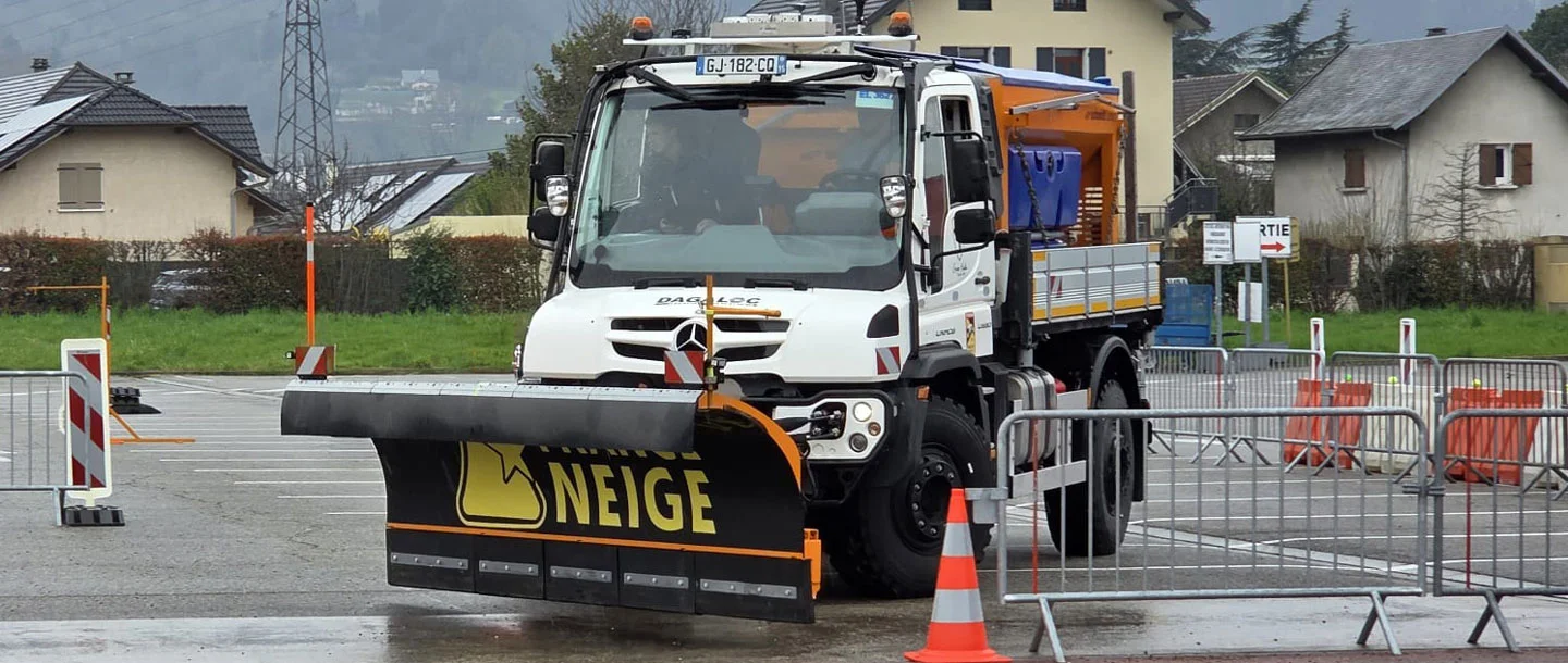 Ein Mercedes-Benz Unimog mit einem Schneepflug steht auf einem Parkplatz, umgeben von Sicherheitsabsperrungen, bereit für Winterdienste.