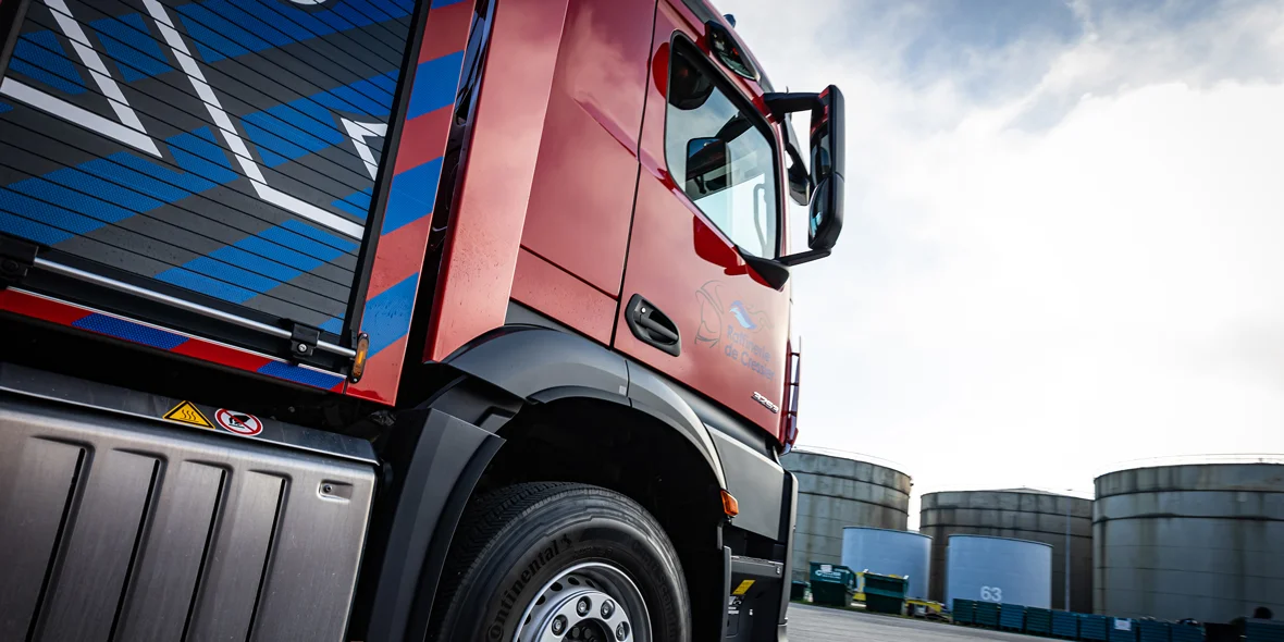 Ein Mercedes-Benz Lkw steht auf einem Gelände mit großen Tanks im Hintergrund. Die Seite des Lkw ist gut sichtbar, einschließlich der Details am Fahrgestell und der Fenster.