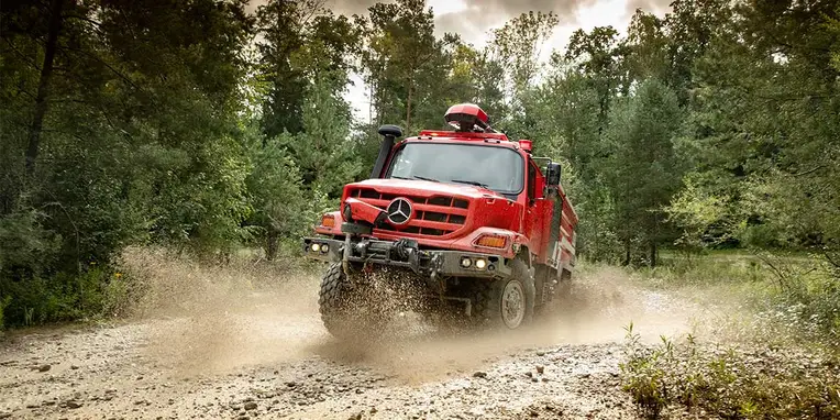 Ein roter Mercedes-Benz Offroad-Fahrzeug fährt durch unwegsames Gelände, wirbelt dabei Staub und Schmutz auf, umgeben von dichtem Wald.