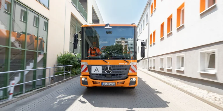 Ein orangefarbener Mercedes-Benz Econic fährt auf einer engen Straße zwischen Gebäuden. Der Fahrer ist sichtbar und trägt eine Arbeitskleidung.