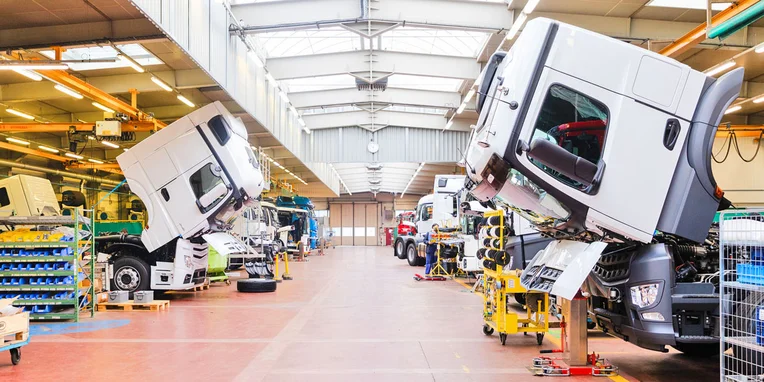 Stabilità e assemblaggio di camion Mercedes-Benz all'interno di un capannone, con cabine sollevate per la lavorazione e diverse attrezzature di montaggio visibili.