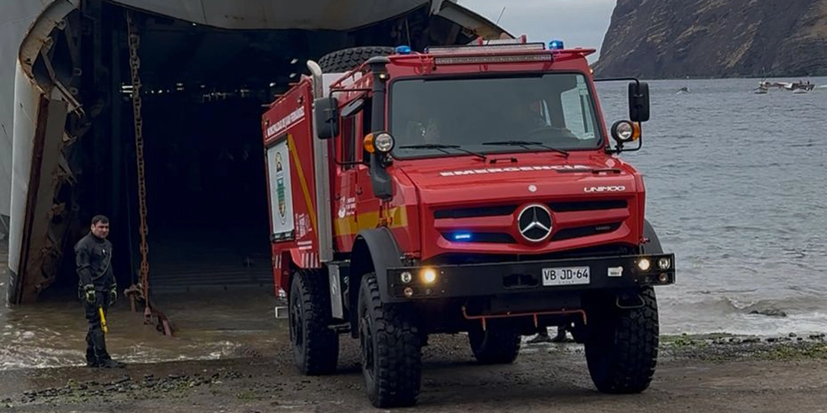 Ein rotes Feuerwehrfahrzeug steht am Strand vor einem großen Schiff. Im Hintergrund sieht man eine Person, die in Schutzkleidung steht. Das Wasser ist ruhig, und im Hintergrund sind einige Boote zu sehen.