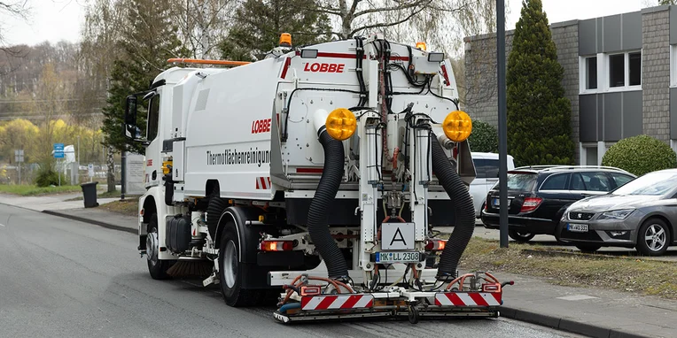 Ein Straßenkehrmaschine der Firma Lobbe steht auf einer Straße. Sie zeigt gelbe Warnleuchten und ist bereit, den Straßenbelag zu reinigen.