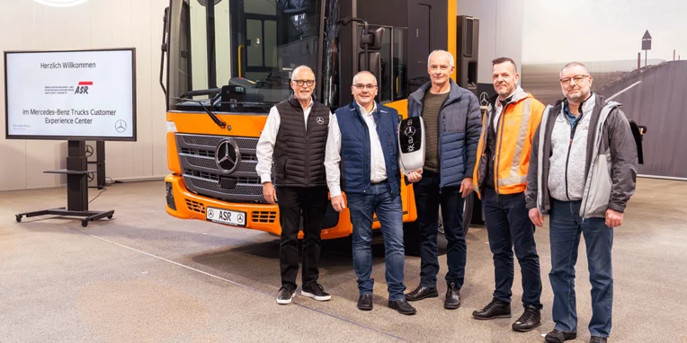 Fünf Personen stehen vor einem orangefarbenen Mercedes-Benz Lkw im Mercedes-Benz Trucks Customer Experience Center, während ein Begrüßungsschild im Hintergrund sichtbar ist.