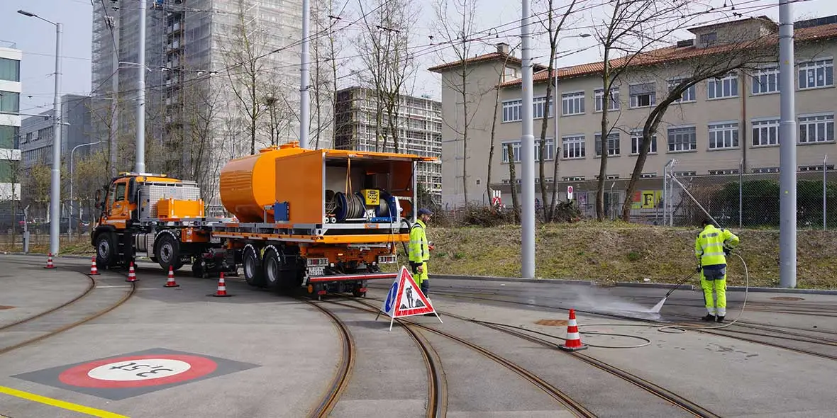 Arbeiter reinigen Gleise mit einem Spezialfahrzeug in einer städtischen Umgebung. Die orangefarbene Maschine ist mit Warnschildern umgeben. Daneben stehen mehrere Verkehrskegel.