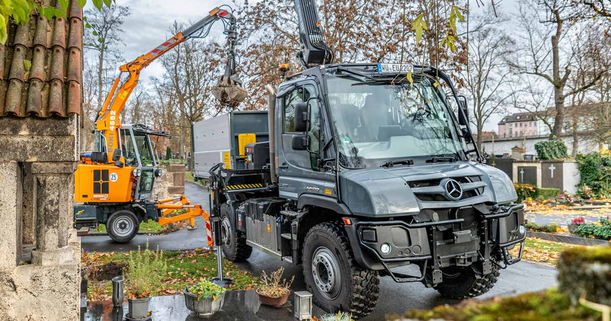 Ein Mercedes-Benz Unimog steht auf einem Friedhof, während ein Kranwagen im Hintergrund Bäume beschneidet. Der Unimog dient als Transportfahrzeug für die Arbeiten.