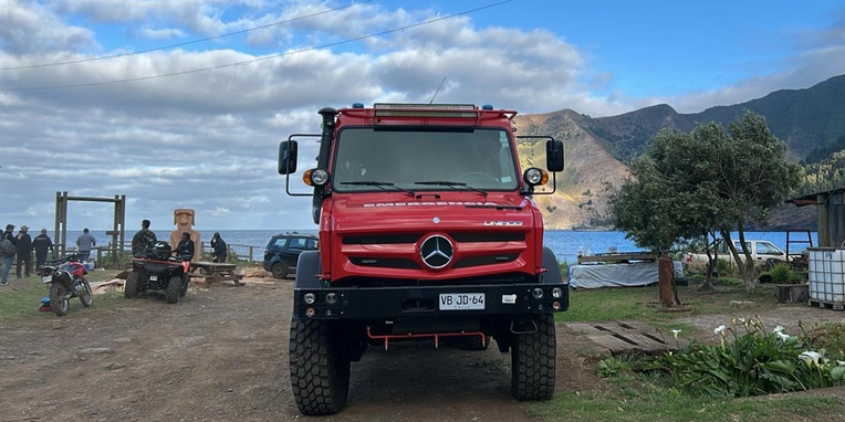 Ein roter Mercedes-Lkw steht auf einem Schotterweg. Im Hintergrund sind Berge und ein Wasserlauf sichtbar. Menschen stehen in Gruppen, während Motorräder in der Nähe geparkt sind. Der Himmel ist bewölkt, was auf wechselhaftes Wetter hinweist.