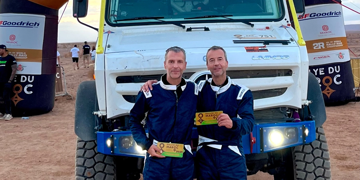 Zwei Fahrer in blauer Fahrerkleidung posieren mit Medaillen vor einem großen Geländewagen von Mercedes-Benz. Im Hintergrund sind Teilnehmer eines Rennens sichtbar.