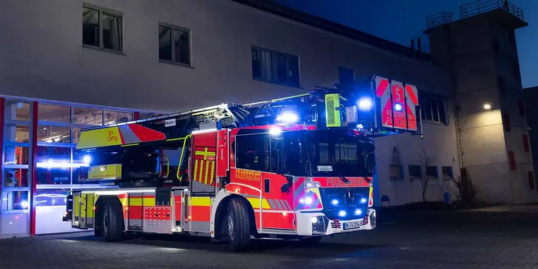 Rettungswagen der Feuerwehr steht bei Nacht vor der Feuerwache mit eingeschaltetem Blaulicht. Die Fahrzeugfront zeigt auffällige Markierungen und eine ausfahrbare Leiter.
