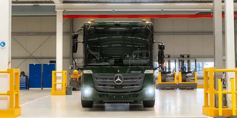 Ein grüner Mercedes-Benz Bus steht in einer modernen Lagerhalle. Die Beleuchtung ist eingeschaltet, und im Hintergrund sind mehrere Gabelstapler und Werkzeuge zu erkennen. Der Fokus liegt auf dem Bus, der zentral im Bild positioniert ist.
