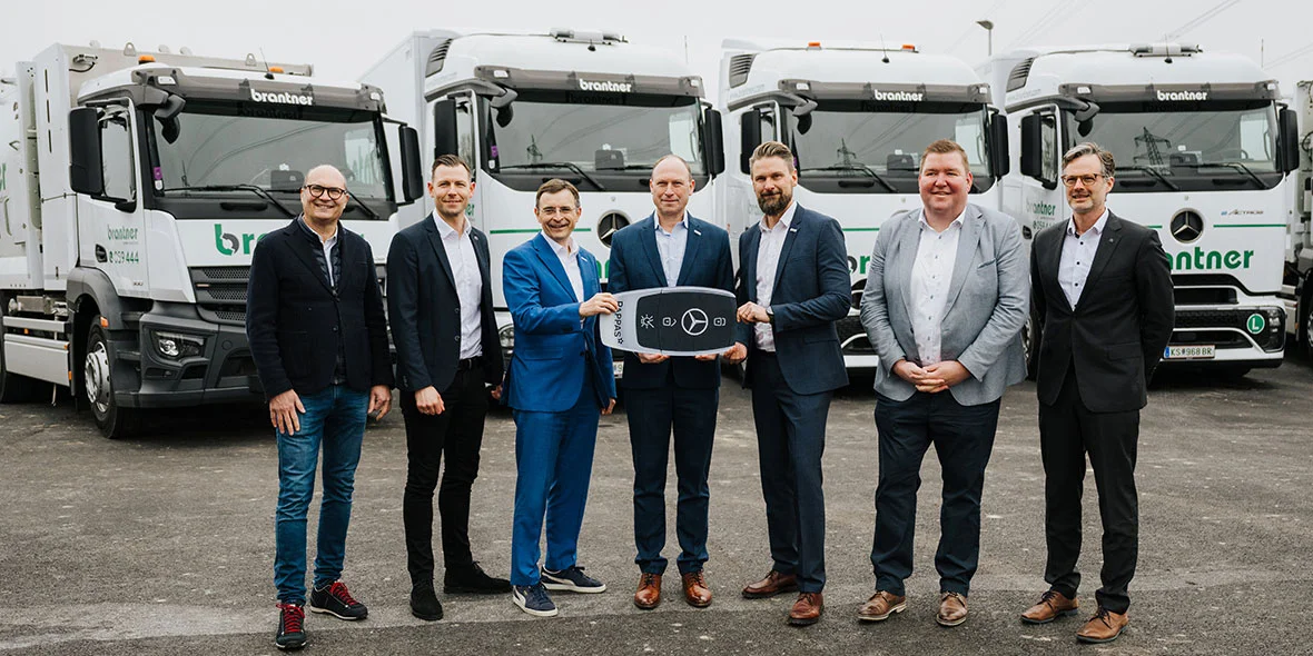 Eine Gruppe von sieben Männern, die im Vordergrund stehen und einen Schlüssel mit dem Mercedes-Benz-Logo halten, während im Hintergrund mehrere Mercedes-Benz-Lkw sichtbar sind.