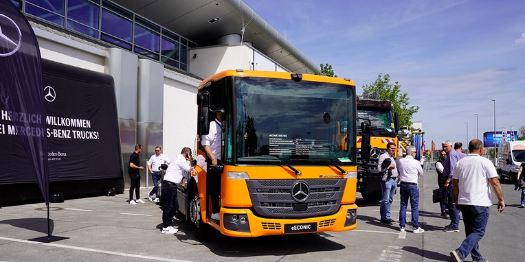 Ein orangefarbenes Mercedes-Benz Nutzfahrzeug steht auf einem Ausstellungsgelände, während Menschen um es herum stehen und das Fahrzeug inspizieren. Im Hintergrund sind weitere Lkw zu sehen.