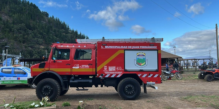 Ein rotes Feuerwehrfahrzeug mit der Aufschrift "Municipalidad de Juan Fernández" steht auf einem Gelände mit Bäumen im Hintergrund. Das Fahrzeug hat große Reifen und ist für schwieriges Gelände ausgelegt. Im Hintergrund sind andere Fahrzeuge und eine offene Struktur zu sehen.