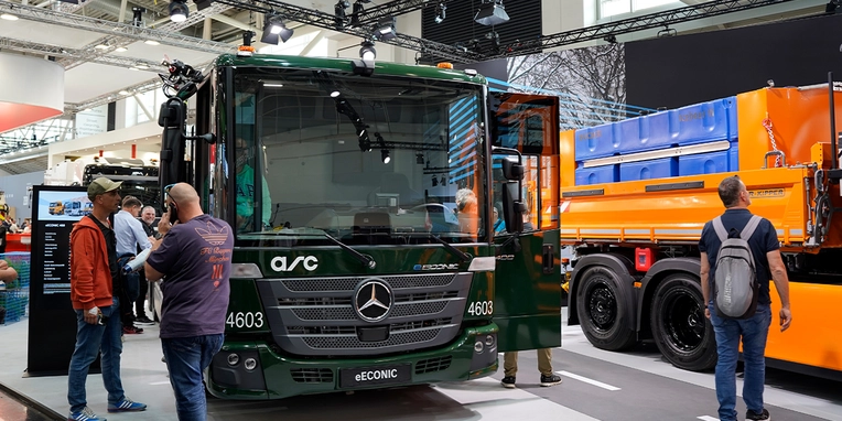 Der Mercedes-Benz eConic, ein moderner Abfallfahrzeug, steht auf einer Messe, während mehrere Personen es betrachten und Informationen austauschen.