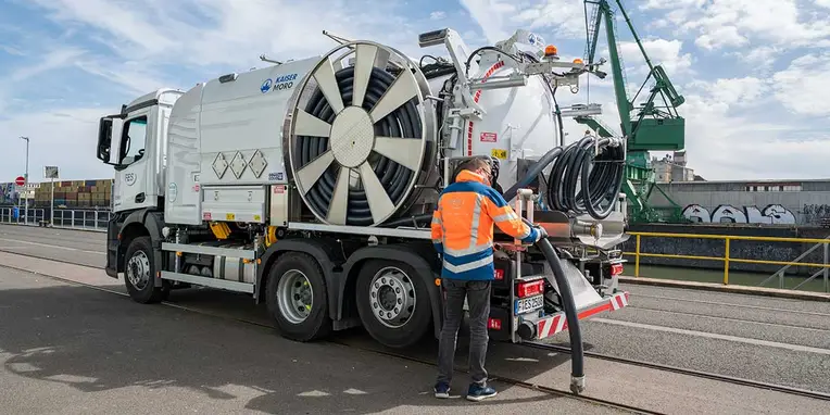 Ein Mercedes-Benz Lkw mit großer Ventilator-Ausrüstung wird von einer Person bedient, die einen Schlauch anschließt. Die Szenerie befindet sich in einem industriellen Hafenbereich.