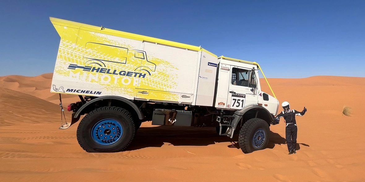 Ein Mercedes-Benz-Lkw steht in einer trockenen Sandlandschaft, während ein Fahrer mit dem Daumen nach oben posiert. Die Fahrzeugseite trägt ein auffälliges gelbes Design mit Sponsorenschrift.