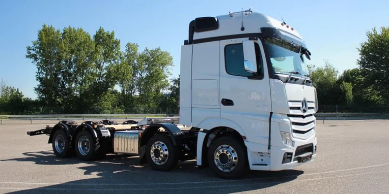 Camion Mercedes-Benz bianco in posizione laterale, con cabina alta e motore visibile. Il veicolo è allestito per il trasporto pesante, in un'area pavimentata con alberi sullo sfondo.