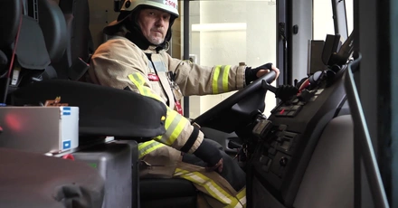 Ein Feuerwehrmann sitzt am Steuer eines Feuerwehrautos, trägt Schutzkleidung und einen Helm. Er schaut ernsthaft nach vorne, bereit für den Einsatz.