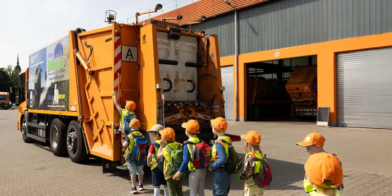 Eine Gruppe von Kindern in orangefarbenen Hüten steht vor einem Mercedes-Benz Econic Müllwagen, der an einer Entsorgungsstation parkt. Kinder tragen Rucksäcke und gelbe Warnwesten.