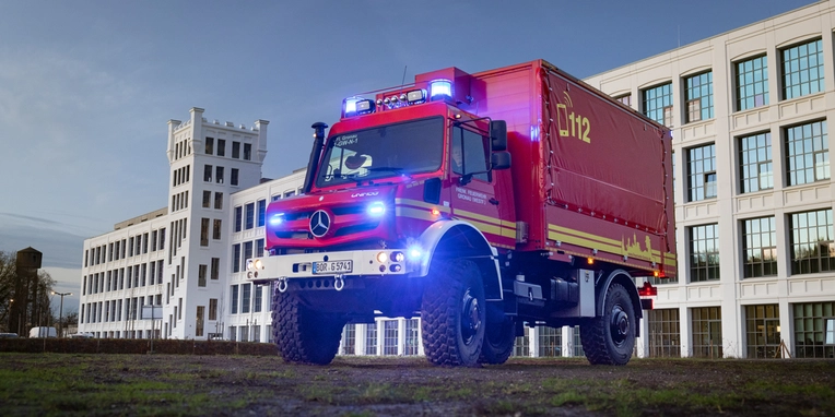 Ein roter Einsatzfahrzeug von Mercedes-Benz, ausgestattet mit Blaulicht und der Aufschrift „112“, steht auf einer Wiese vor einem modernen Gebäude.