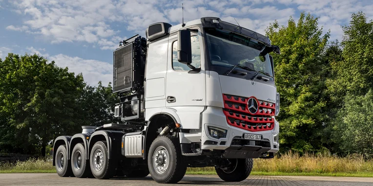 Camion Mercedes-Benz con cabine e rimorchio, di colore bianco, parcheggiato su una superficie asfaltata, circondato da alberi verdi.