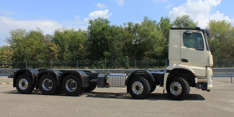 Camion Mercedes-Benz bianco con cabina alta, visibile lateralmente, dotato di quattro assi e ruote in metallo, su una superficie stradale con vegetazione sullo sfondo.