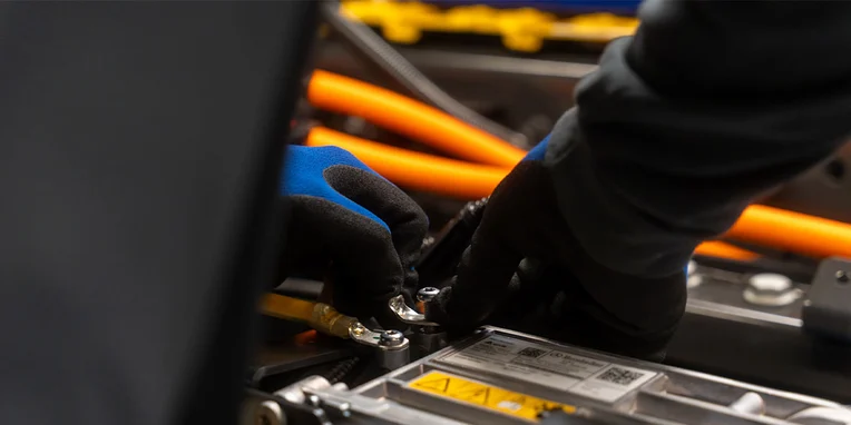 Ein Mechaniker trägt schwarze Handschuhe und arbeitet an einem Batteriemodul. Er zieht eine Verbindung an, während orangefarbene Kabel im Hintergrund sichtbar sind. Die Umgebung deutet auf eine technische Werkstatt hin.
