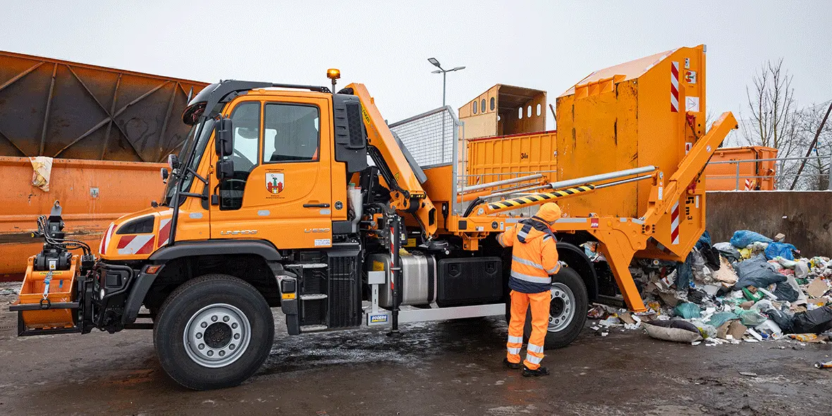 Ein Mercedes-Benz Müllfahrzeug mit einem orangefarbenen Aufbau entleert Müll in einer Recyclingstation, während der Fahrer in einer leuchtenden Warnjacke arbeitet.