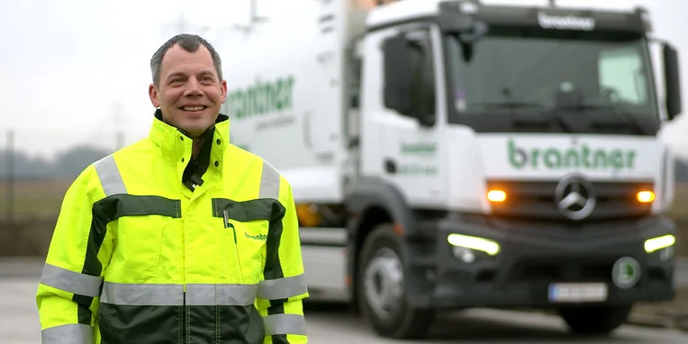 Ein lachender Mann in einem reflektierenden gelben Arbeitsanzug steht vor einem Mercedes-Benz Lkw mit dem Logo der Firma Brantner.