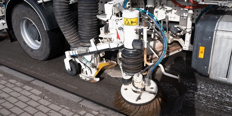 Eine Reinigungseinheit mit Besen und Schläuchen, die auf einer Straßenoberfläche arbeitet. Die Mechanik gehört zu einem Fahrzeug, das für die Straßenreinigung konzipiert ist.