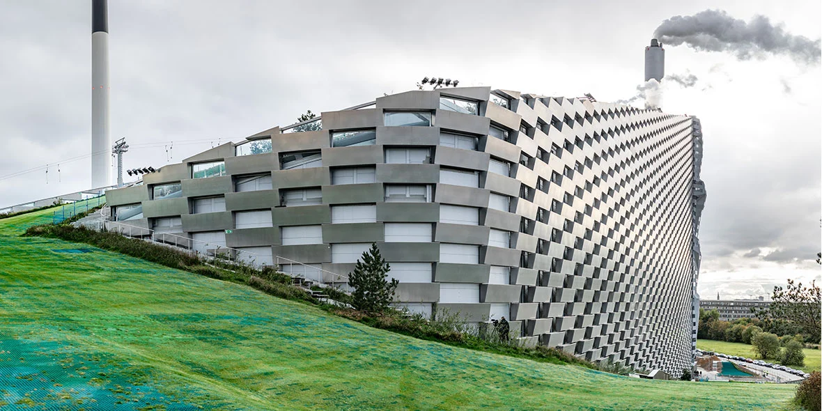 Modernes Gebäude mit einer gewellten Fassade, umgeben von Rasenflächen. Im Hintergrund ist ein Schornstein sichtbar, aus dem Rauch aufsteigt. Standort ist anscheinend eine Sport- oder Freizeitstätte.