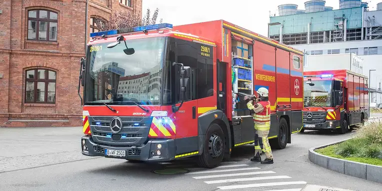 Einsatzfahrzeug der Feuerwehr von Mercedes-Benz steht an einer Kreuzung, während ein Feuerwehrmann in Schutzkleidung sich auf den Einsatz vorbereitet.