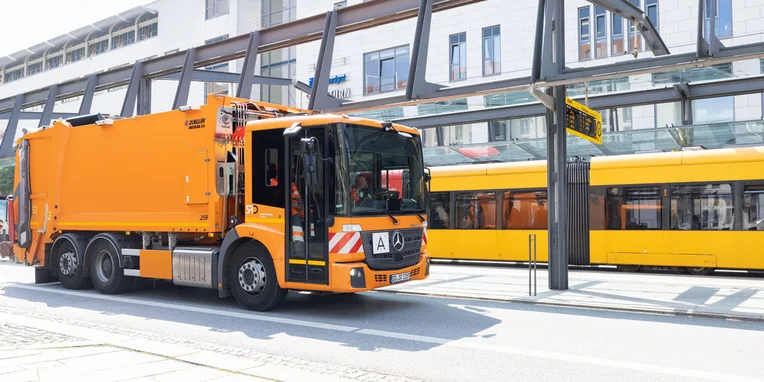 Ein orangefarbener Mercedes-Benz Econic Müllwagen hält an einer Haltestelle neben einer gelben Straßenbahn.