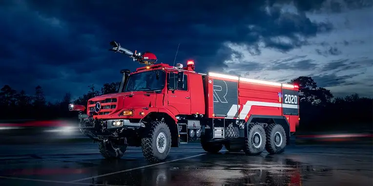 Ein roter Mercedes-Benz Feuerwehrauto steht bei Nacht auf einer regnerischen Straße, beleuchtet von Scheinwerfern und mit einem großen Löschmonitor auf dem Dach.