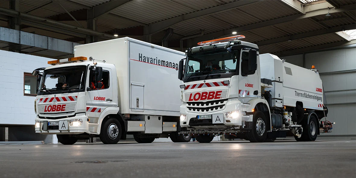 Zwei Mercedes-Benz Fahrzeuge stehen in einer Fahrzeughalle. Links ein weißer Lkw mit Havarie-Management-Anhänger, rechts ein Spezialfahrzeug mit der Aufschrift „LOBBE“.