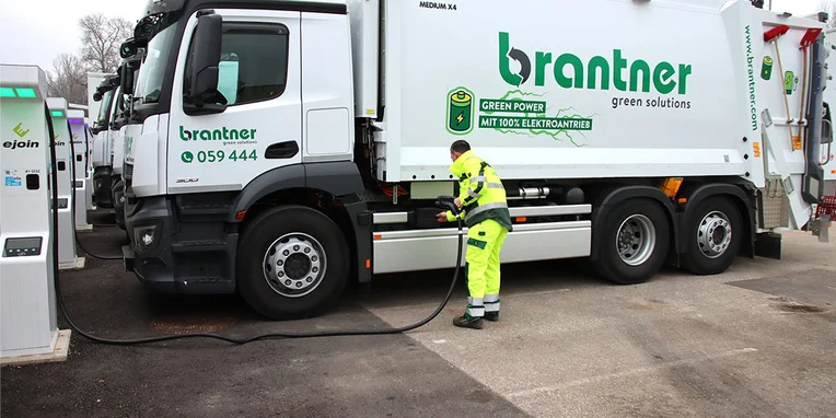 Ein Mitarbeiter in einheitlicher Arbeitskleidung tankt einen Elektro-Abfallwagen der Marke Brantner an einer Ladestation, während mehrere Fahrzeuge im Hintergrund parken.