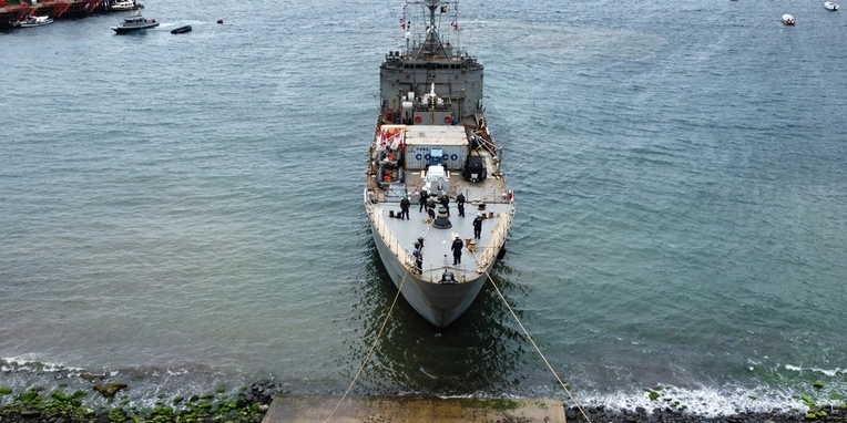 Ein Schiff liegt im Wasser, mit mehreren Personen an Deck. Im Hintergrund sind andere Boote und landseitige Elemente sichtbar. Das Wasser ist ruhig und die Szene vermittelt ein maritimes Ambiente.