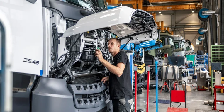 Un tecnico che lavora su un camion Mercedes-Benz, sollevando il cofano e controllando il motore in un ambiente di produzione, circondato da attrezzature e altri veicoli in fase di assemblaggio.