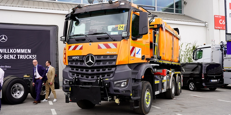 Ein orangefarbener Mercedes-Benz Arocs Lkw steht im Vordergrund, während im Hintergrund Personen und weitere Nutzfahrzeuge zu sehen sind. Der Lkw ist für den Transport von Baustellenmaterialien ausgelegt.