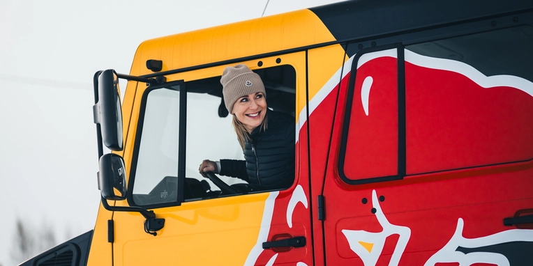 Eine Frau mit Mütze lächelt fröhlich aus dem Fenster eines roten und gelben Lastwagens mit Red Bull-Design. Der Lastwagen steht in einer verschneiten Umgebung.