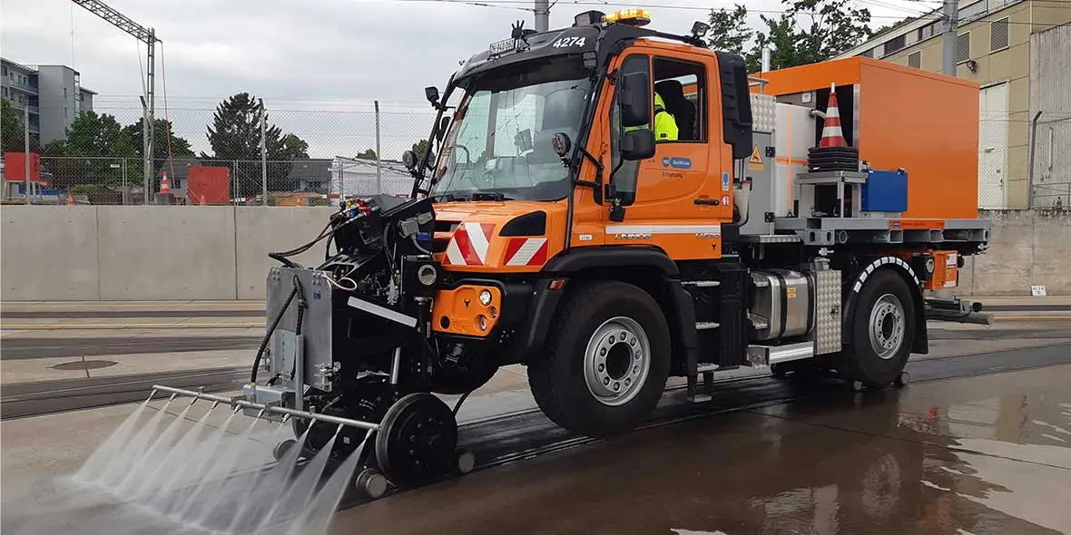Ein oranger Mercedes-Benz Straßenreinigungsfahrzeug mit Wasseranwendung steht auf einer nassen Oberfläche, ausgestattet mit einer Kehrausrüstung und einem speziellen Reinigungssystem.